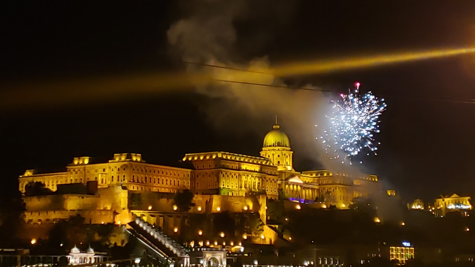 Buda Castle glowing at night as fireworks break open over the hill