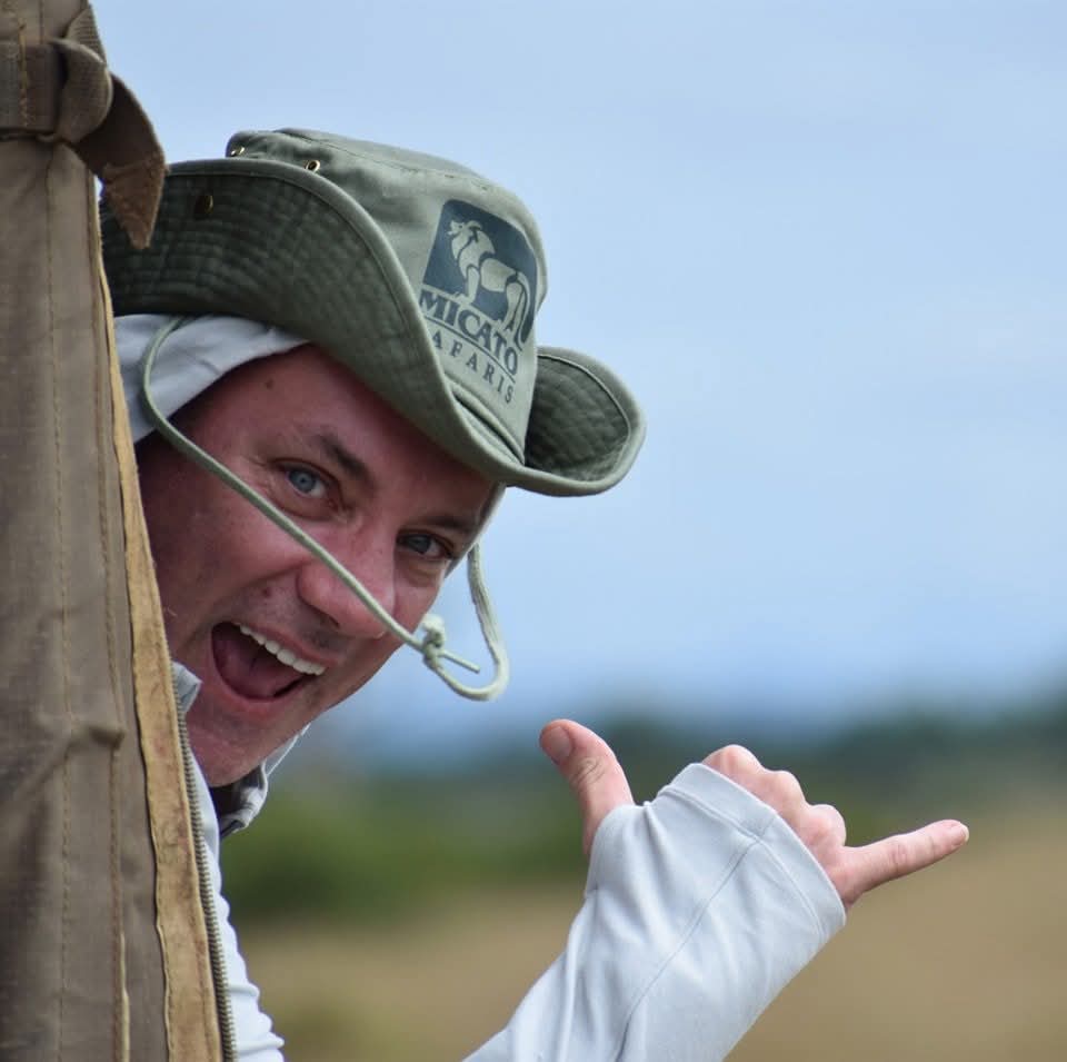 Scott Barlow on safari in Kenya looking happy leaning out of a vehicle in front of a tent, wearing a safari hat