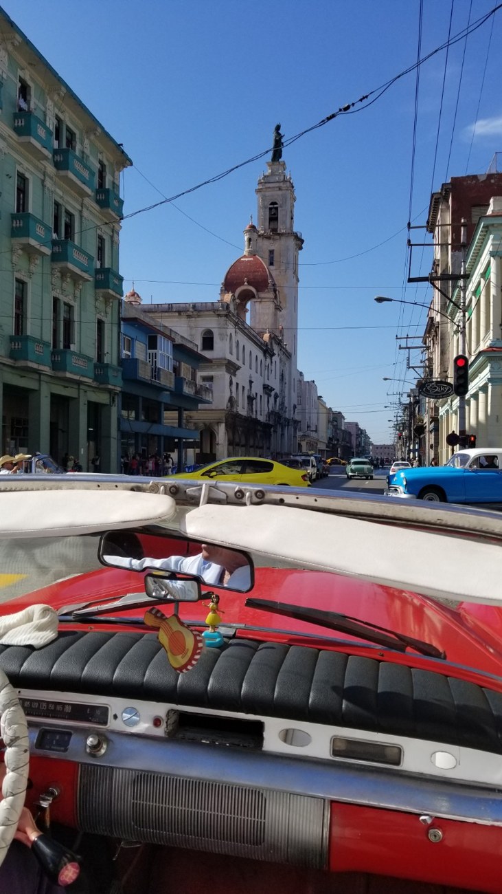 View forward from inside a red convertible riding through Havana toward a church tower