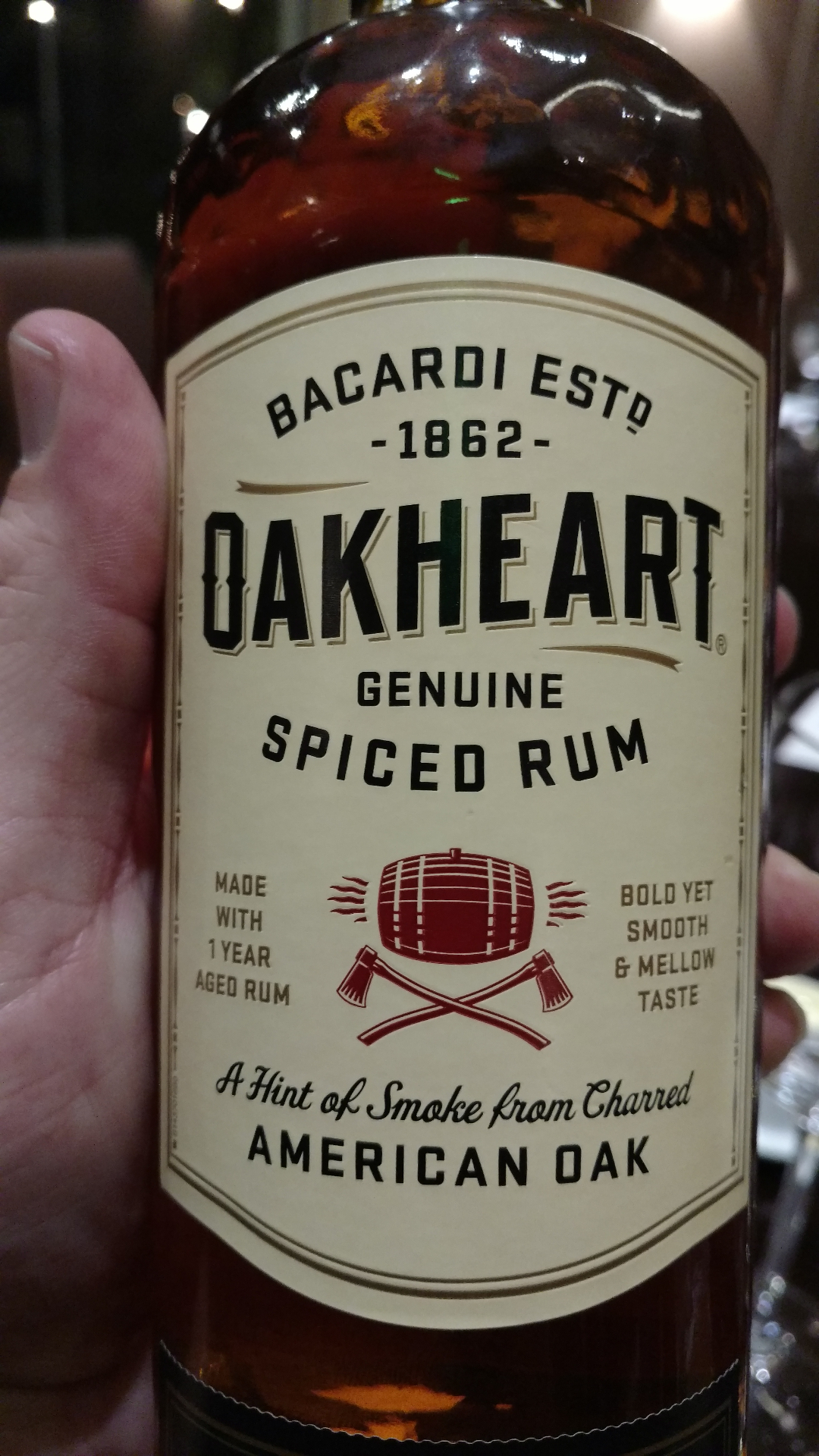 A bottle of Bacardi Oakheart Genuine Spiced Rum held in hand, cream label with a red barrel and crossed axes.