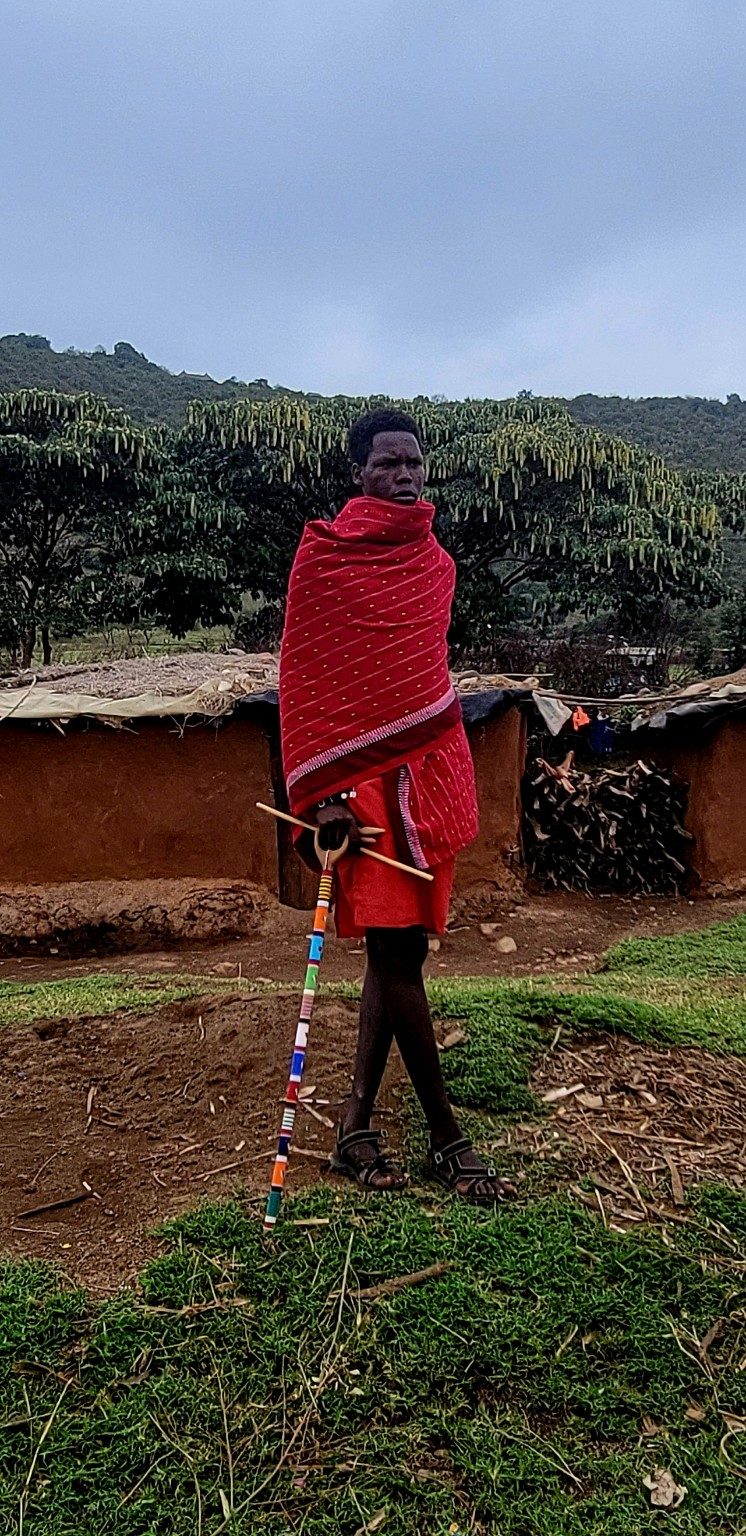 Maasai man in a red shuka holding a beaded staff outside a mud-walled home in Narok County