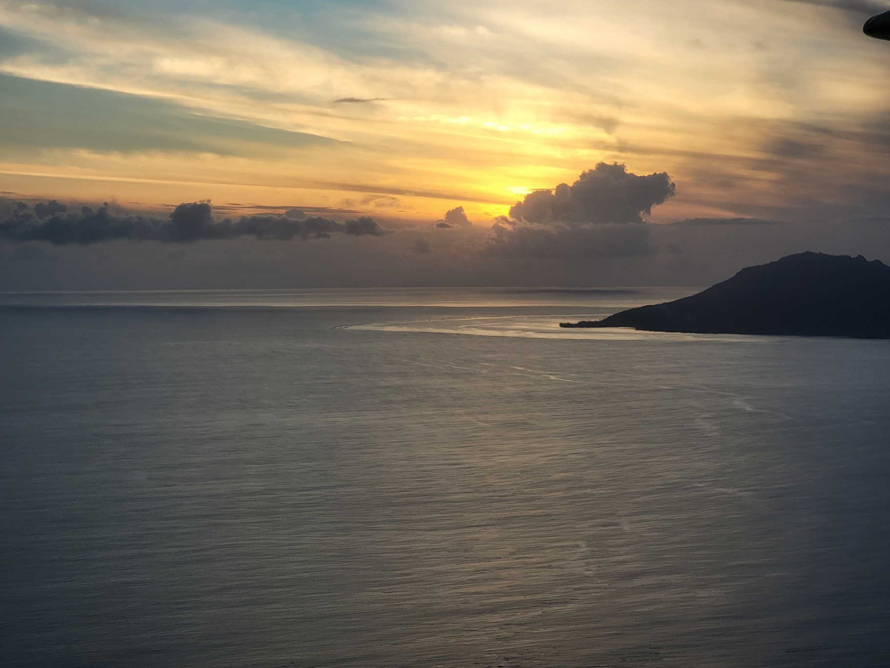 Aerial view from the plane window at sunset showing a Tahitian island silhouette and reef passage