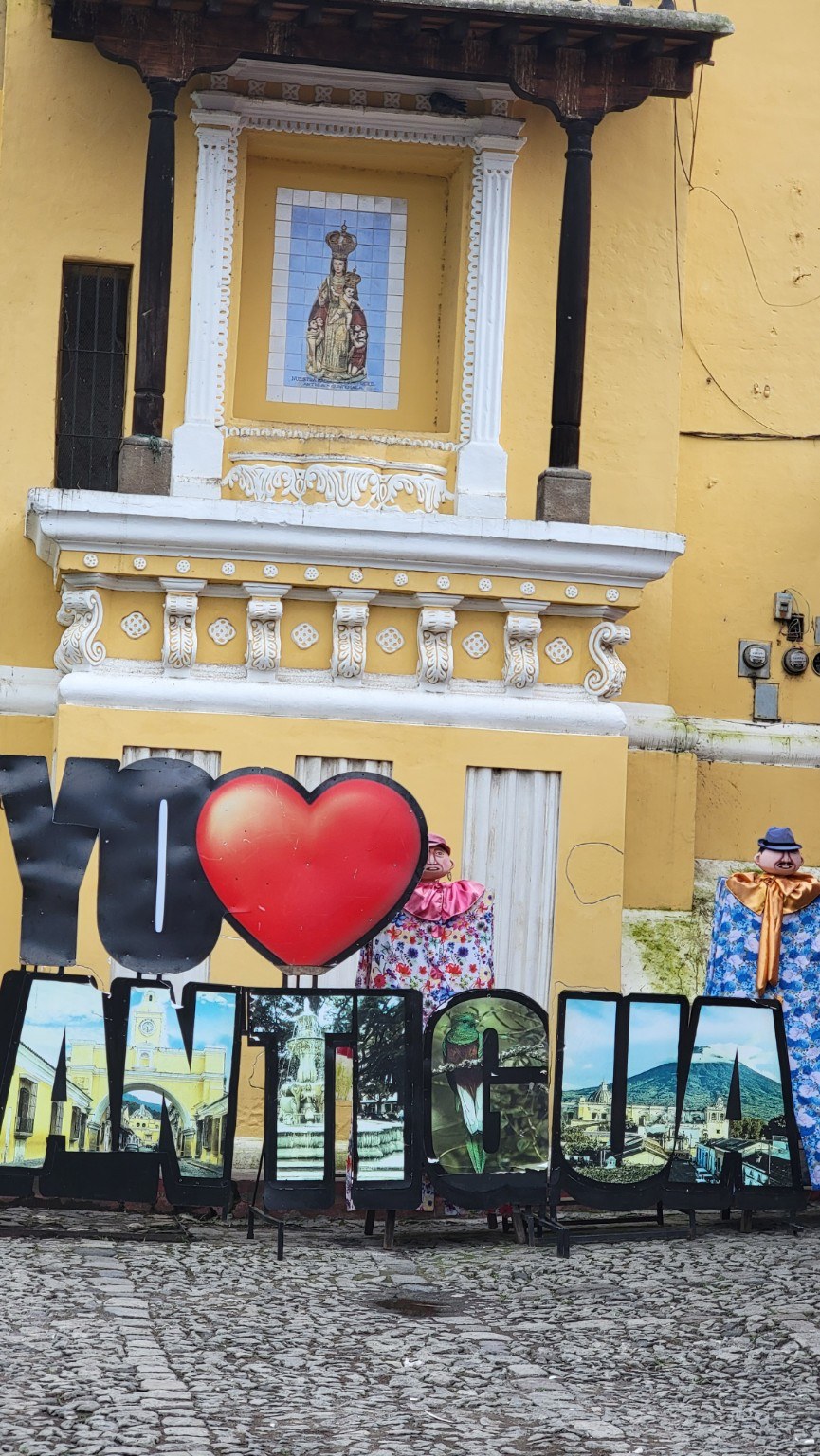 Large 'Yo amo Antigua' letters with marionette figures in front of a yellow colonial wall