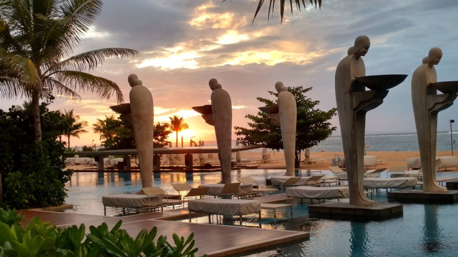 Sunset over a Bali resort pool with sculptural figures rising from the water