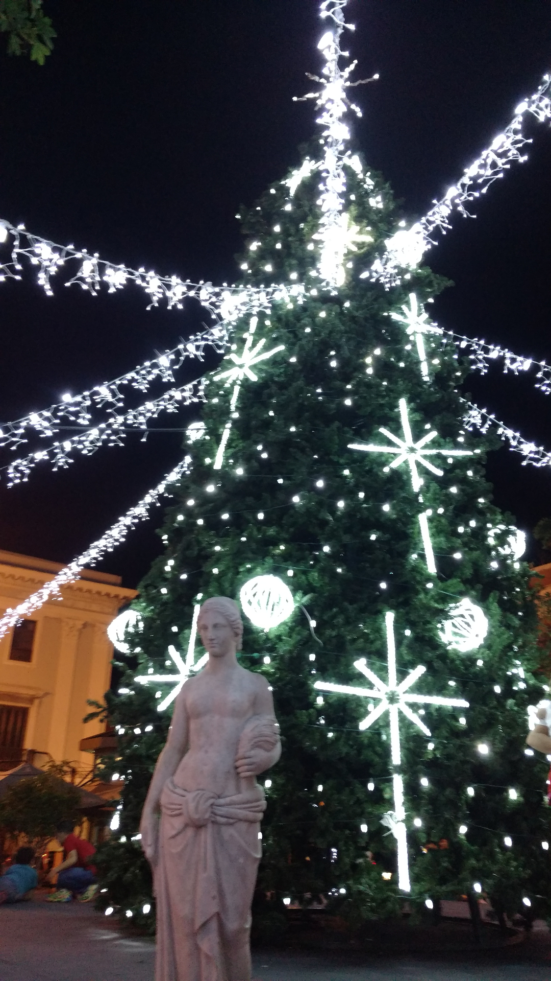 A tall Christmas tree strung with white lights in an Old San Juan plaza, radiating light garlands over a classical statue in the foreground.