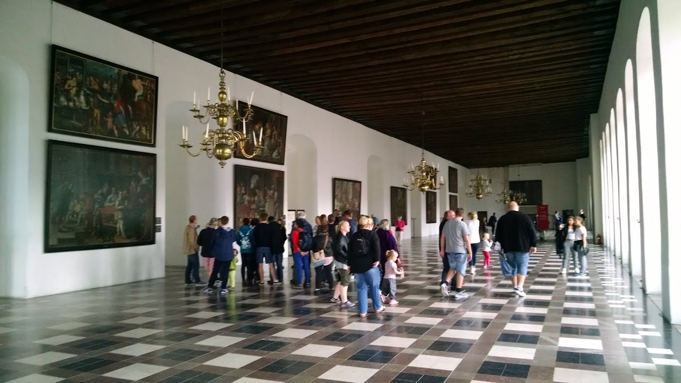 A long interior hall at Kronborg, with checkerboard floors and portraits watching the tourists pass by